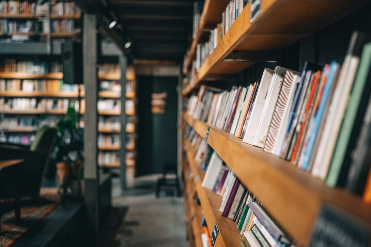 A close-up view of bookshelves filled with books in a library or bookstore, with a blurred reading area visible in the background.